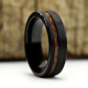 May include: A black ring with a brushed metal finish and a wood inlay. The ring has a dark, sleek appearance with a contrasting light brown wood grain center. The ring's design is modern and minimalist.