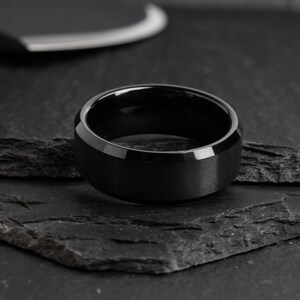 May include: A black ring with a matte finish and beveled edges. The ring is set on a dark, textured surface. The ring's design is simple and modern, suitable for everyday wear.