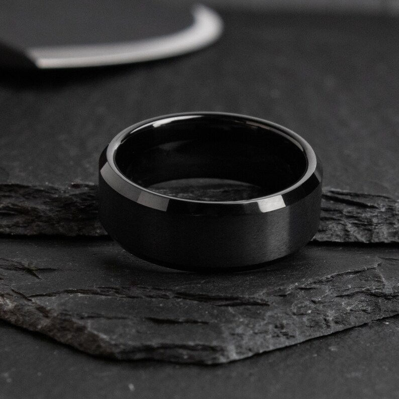 May include: A black ring with a matte finish and beveled edges. The ring is set on a dark, textured surface. The ring's design is simple and modern, suitable for everyday wear.