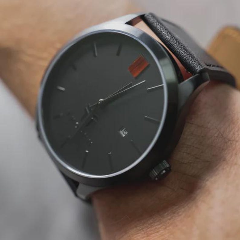 May include: A close-up of a black wristwatch with a black leather band. The watch face is black with silver hands and markers. A small orange accent is visible on the watch face. The watch is on a wrist.