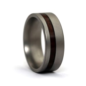 Puede incluir: Un anillo plateado con una incrustación de madera marrón oscuro. El anillo tiene un perfil plano y un acabado liso y mate. La incrustación de madera recorre el centro del anillo. Un diseño simple y elegante.