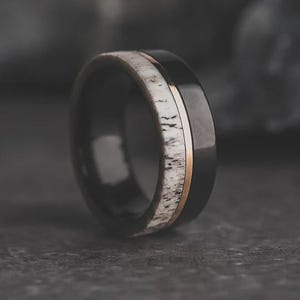 May include: A black ring with a gold band and a white and brown antler inlay. The ring is made of a dark, shiny material and is set against a dark background. The ring is a close-up shot.
