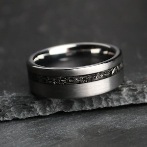 May include: A dark grey ring with a textured black inlay. The ring has a brushed metal finish and a wide band. The inlay appears to be made of a dark, possibly stone-like material.