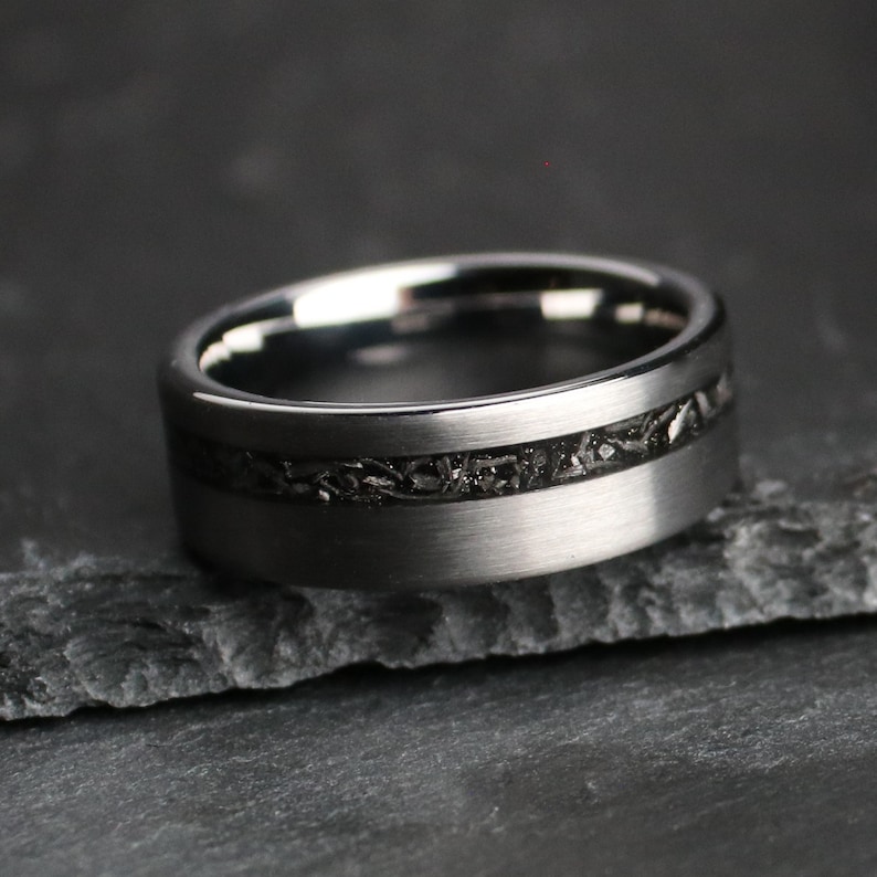 May include: A dark grey ring with a textured black inlay. The ring has a brushed metal finish and a wide band. The inlay appears to be made of a dark, possibly stone-like material.