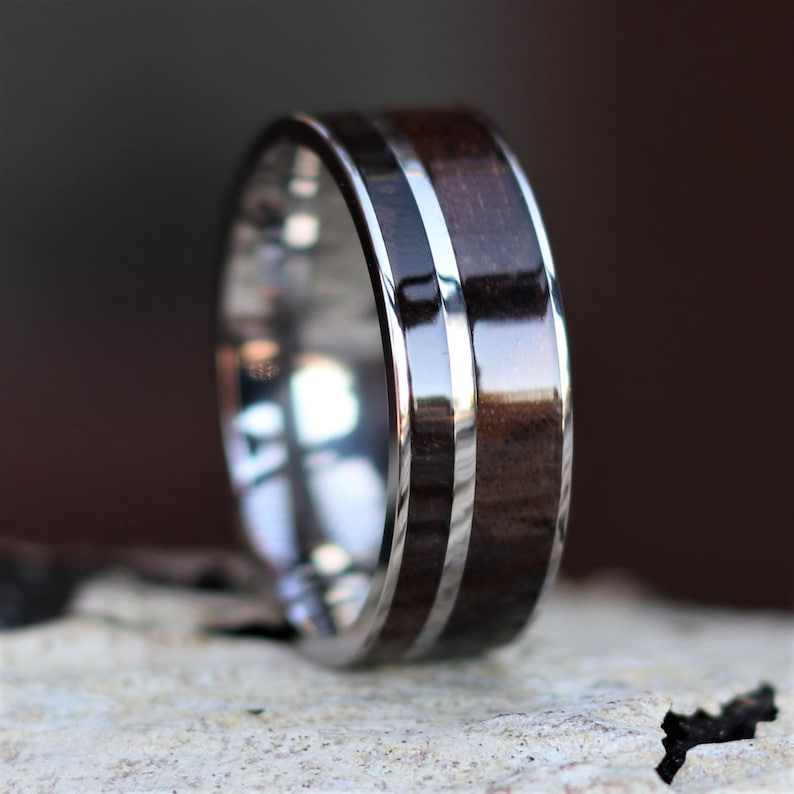 May include: A close-up of a ring with a polished silver band and a dark brown wood inlay. The wood is a rich, warm color, and the silver band has a reflective surface. The ring is a simple, elegant design.