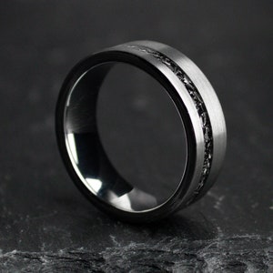May include: A silver ring with a black inlay. The ring features a brushed metal finish on the outer band and a dark, textured inlay around the centre. The ring is presented on a dark, textured surface.