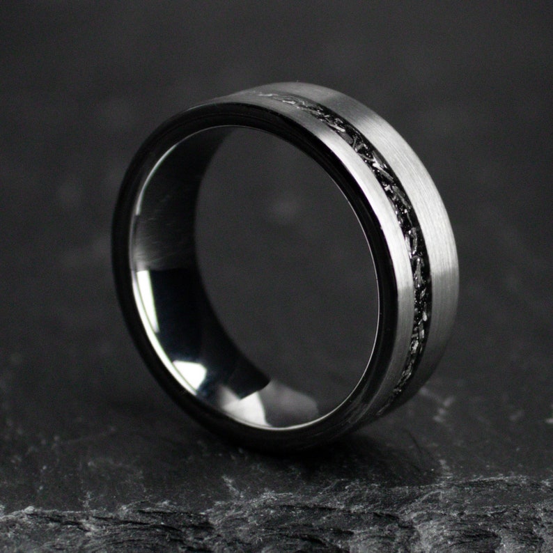 May include: A silver ring with a black inlay. The ring features a brushed metal finish on the outer band and a dark, textured inlay around the centre. The ring is presented on a dark, textured surface.