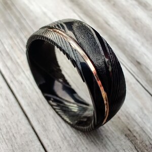 May include: A black ring with a textured surface and a thin, copper-colored stripe. The ring has a unique, patterned design and a polished finish. The ring is a piece of jewelry.