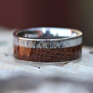 May include: A silver ring with a wide band. The ring features a center section of dark brown wood with a textured, light-colored band above and below. The ring is set against a blurred background.