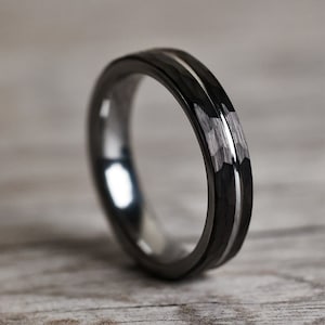 Pu&ograve; includere: Un anello nero con intarsio argentato. L'anello ha una superficie lucida e un motivo geometrico strutturato sulla fascia esterna. L'anello &egrave; posto su uno sfondo chiaro con venature del legno.