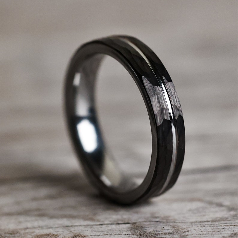 Pu&ograve; includere: Un anello nero con intarsio argentato. L'anello ha una superficie lucida e un motivo geometrico strutturato sulla fascia esterna. L'anello &egrave; posto su uno sfondo chiaro con venature del legno.