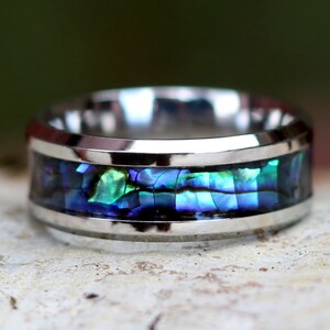 May include: A stainless steel ring with a band of iridescent abalone shell. The abalone shell displays shades of blue, green, and purple. The ring has a polished, reflective finish, and the abalone is set between two silver bands.