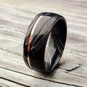 May include: A black Damascus steel ring with a copper inlay. The ring has a textured pattern and a polished finish. The copper inlay runs down the center of the ring. The ring is on a wooden surface.