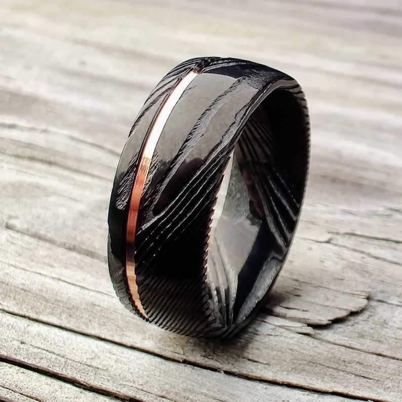May include: A black Damascus steel ring with a copper inlay. The ring has a textured pattern and a polished finish. The copper inlay runs down the center of the ring. The ring is on a wooden surface.