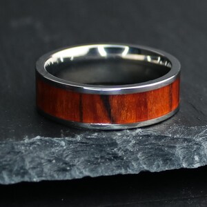 May include: A silver ring with a wide band of reddish-brown wood grain. The ring has a polished silver interior and exterior edges. The wood grain has dark brown streaks. The ring is displayed on a dark gray surface.