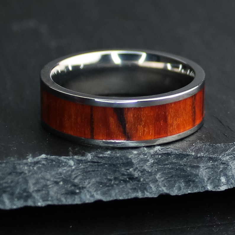 May include: A silver ring with a wide band of reddish-brown wood grain. The ring has a polished silver interior and exterior edges. The wood grain has dark brown streaks. The ring is displayed on a dark gray surface.