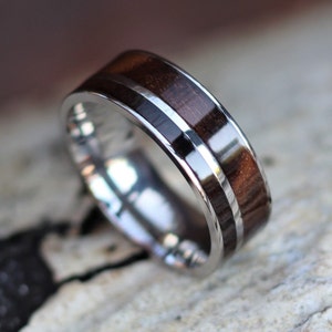May include: A close-up of a silver ring with a dark brown wood inlay. The ring has a polished, reflective surface and a thin silver band bordering the wood. The wood grain is visible, and the ring appears to be handcrafted.