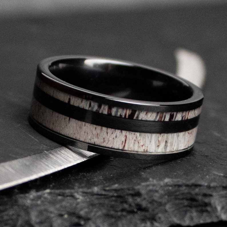 Puede incluir: Un anillo negro con una banda de material de asta de color marr&oacute;n claro y tonos rojizos, enmarcado por dos bandas de metal negro. El anillo tiene un exterior negro pulido y un dise&ntilde;o moderno.