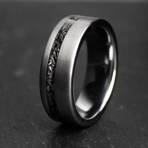 May include: A silver-coloured ring with a black inlay, shown in close-up. The ring has a brushed metal finish and a dark, textured central band. The ring is a wide band style, with a sleek, modern design.