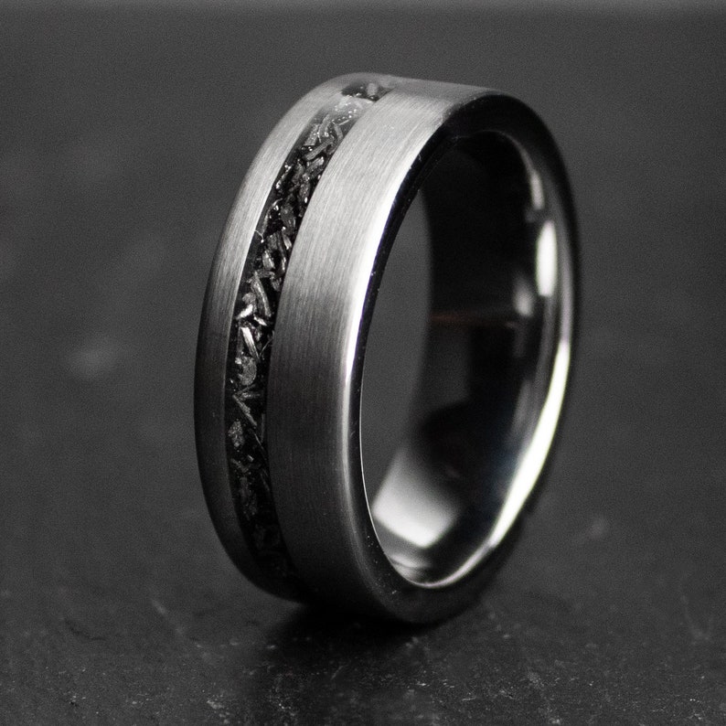 May include: A silver-coloured ring with a black inlay, shown in close-up. The ring has a brushed metal finish and a dark, textured central band. The ring is a wide band style, with a sleek, modern design.