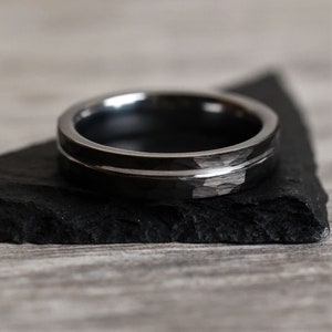 Pu&ograve; includere: Un anello grigio scuro con una texture martellata e una sottile fascia argentata lucida. L'anello &egrave; appoggiato su una superficie nera strutturata. Il design dell'anello &egrave; semplice e moderno, adatto all'uso quotidiano.