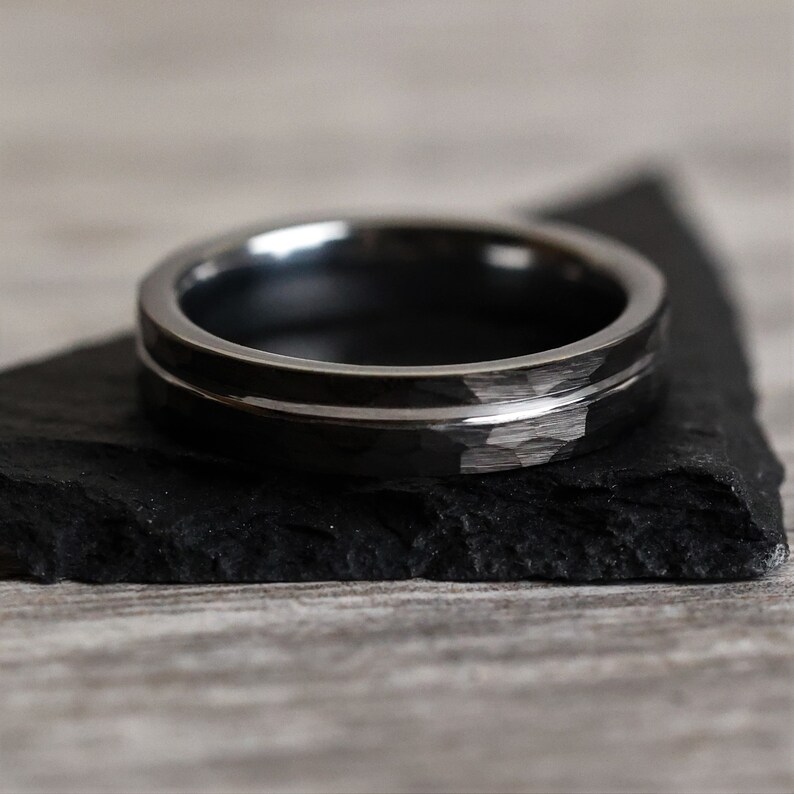 Pu&ograve; includere: Un anello grigio scuro con una texture martellata e una sottile fascia argentata lucida. L'anello &egrave; appoggiato su una superficie nera strutturata. Il design dell'anello &egrave; semplice e moderno, adatto all'uso quotidiano.