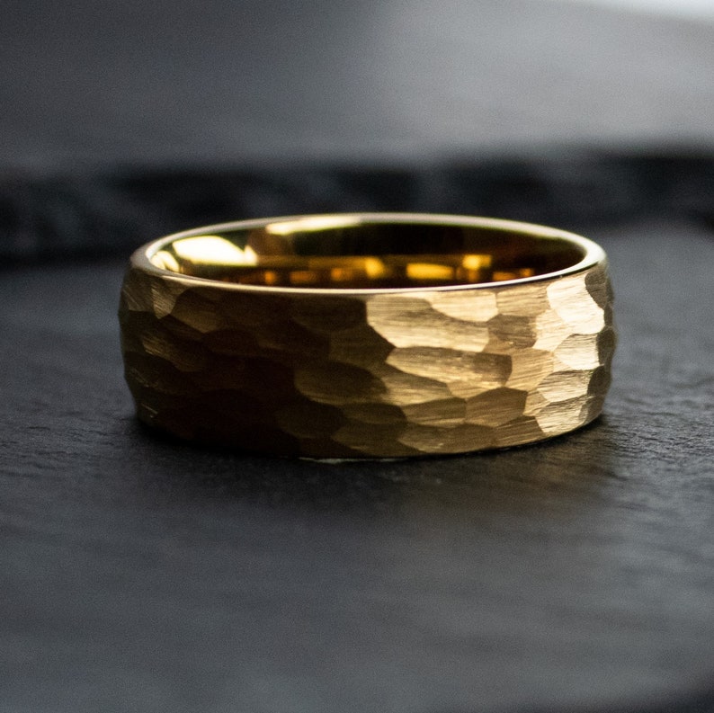 May include: A gold-colored ring with a hammered texture. The ring has a wide band and a matte finish, with a slightly rounded interior. The ring is set against a dark background, highlighting its metallic sheen. Jewelry.