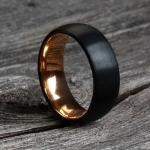 May include: A black ring with a brushed finish and a gold-colored interior. The ring is set against a weathered, gray wooden surface, highlighting its contrasting textures and colors. The ring's design is simple and modern.