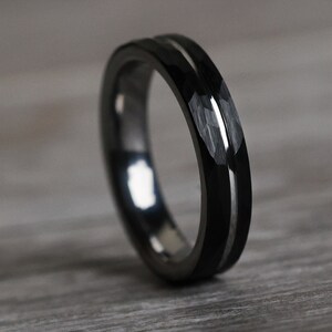 Pu&ograve; includere: Un anello nero con intarsio argentato. L'anello presenta un design sfaccettato sulla fascia esterna, creando un aspetto strutturato. L'anello &egrave; appoggiato su una superficie di legno.