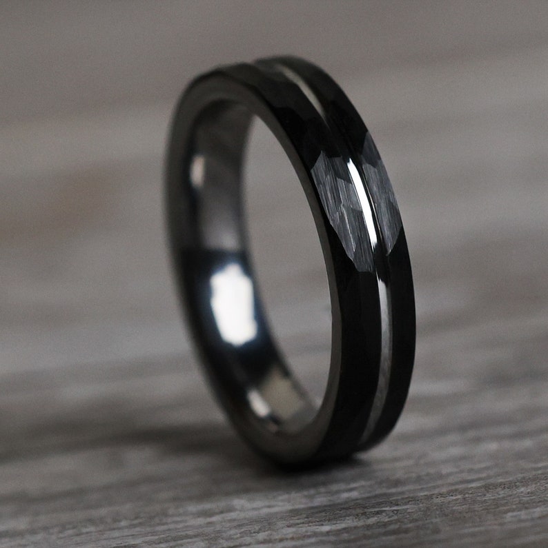 Pu&ograve; includere: Un anello nero con intarsio argentato. L'anello presenta un design sfaccettato sulla fascia esterna, creando un aspetto strutturato. L'anello &egrave; appoggiato su una superficie di legno.