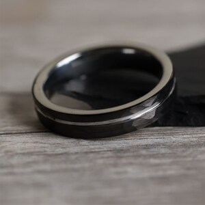 Pu&ograve; includere: Primo piano di un anello nero con una fascia argentata e una superficie strutturata. L'anello ha un design moderno. L'anello &egrave; probabilmente realizzato in tungsteno o fibra di carbonio. L'anello &egrave; su una superficie di legno. Il diametro dell'anello &egrave; di circa 2 cm.