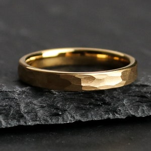 May include: A gold-colored ring with a hammered texture. The ring is set against a dark, textured background. The ring's surface has a matte finish, and the band is approximately 0.1 inches wide.