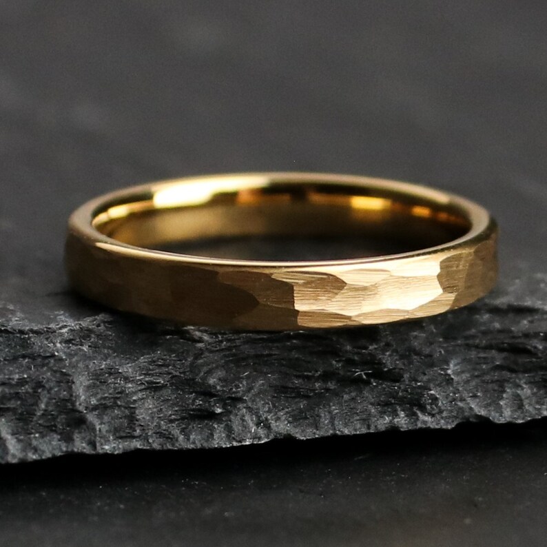 May include: A gold-colored ring with a hammered texture. The ring is set against a dark, textured background. The ring's surface has a matte finish, and the band is approximately 0.1 inches wide.