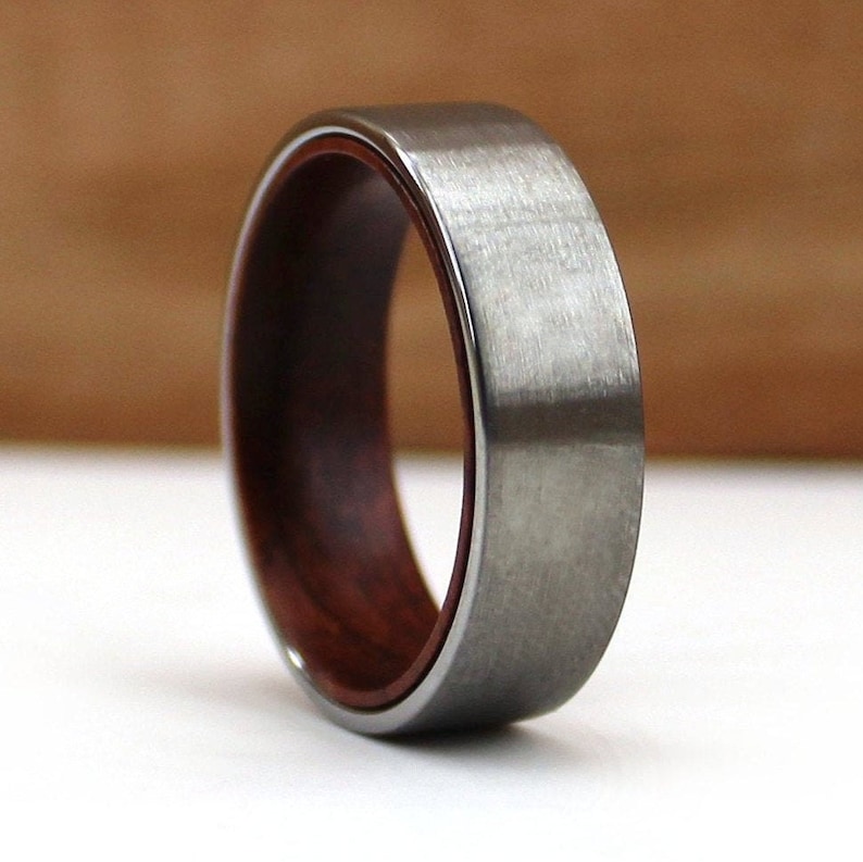 May include: A close-up of a ring with a brushed metal exterior and a dark wood interior. The ring has a flat profile and a polished metal edge. The wood interior is a rich, reddish-brown color.