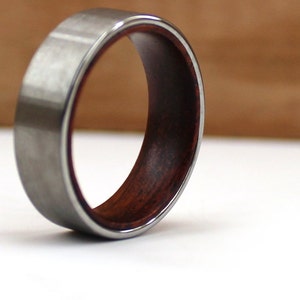 May include: A close-up of a ring with a silver-colored exterior and a dark reddish-brown wooden interior. The ring is set against a white surface with a blurred brown background.