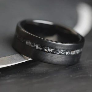 May include: A close-up of a black ring with a textured finish. The ring features a band of light-colored, speckled material inlaid in the center. The ring is set against a dark background, highlighting its design.