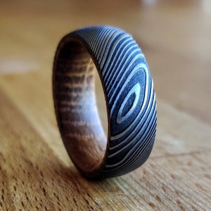 May include: A close-up of a Damascus steel ring with a unique pattern. The ring features a dark, textured exterior with a swirling design and a lighter-colored interior. The ring is displayed on a wooden surface.