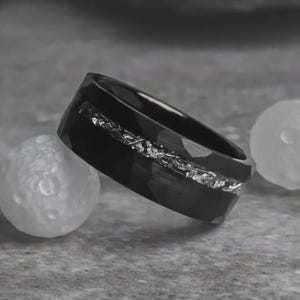 May include: A black ring with a faceted design and a silver inlay. The ring is set against a gray background with two white, spherical objects. The ring's band is approximately 0.3 inches wide.
