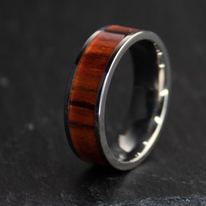 May include: A close-up of a ring with a dark metal band and a wood inlay. The wood is a rich reddish-brown color with dark grain patterns. The ring is set against a dark background.