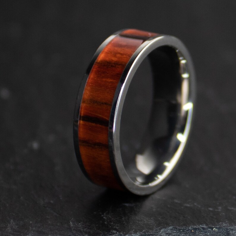 May include: A close-up of a ring with a dark metal band and a wood inlay. The wood is a rich reddish-brown color with dark grain patterns. The ring is set against a dark background.