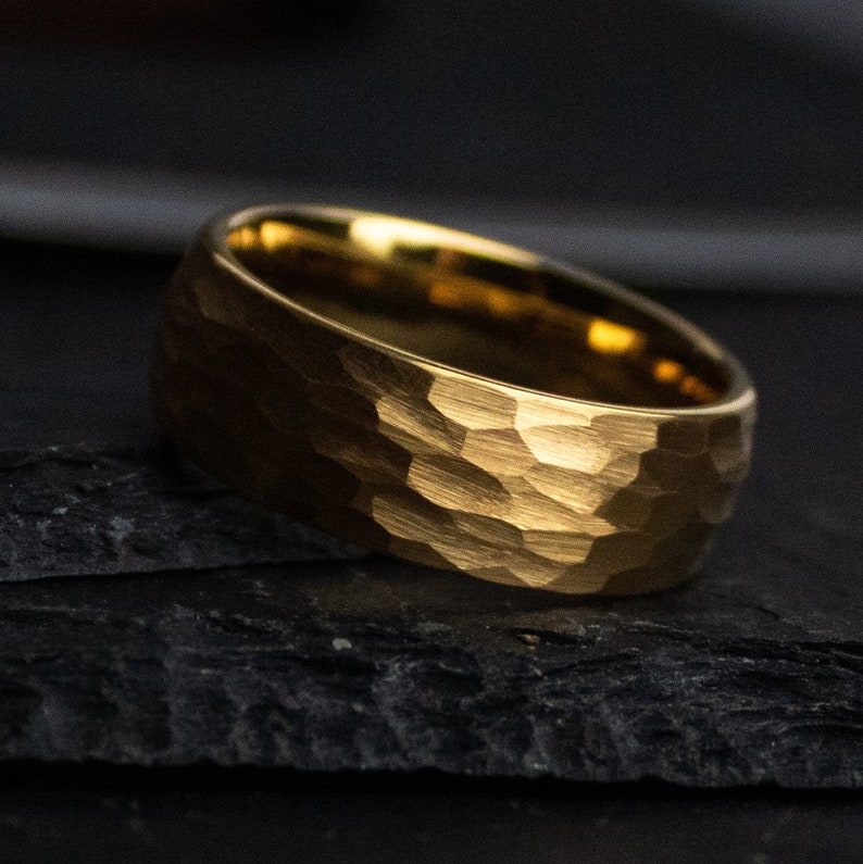 May include: A gold-colored ring with a textured, hammered finish. The ring has a wide band and a rounded shape. The surface of the ring reflects light, creating highlights and shadows. The ring is a piece of jewelry.