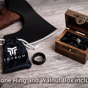 May include: A black silicone ring and a wooden ring box with a ring inside. The ring box is open, revealing a black ring with a silver band. A black bag with the word "THORUM" is next to the ring. An axe is in the background.