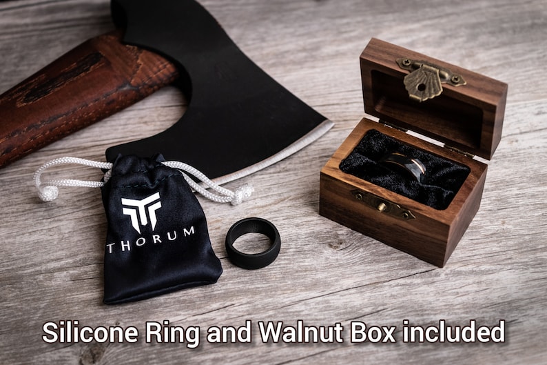 May include: A black silicone ring and a wooden ring box with a ring inside. The ring box is open, revealing a black ring with a silver band. A black bag with the word "THORUM" is next to the ring. An axe is in the background.