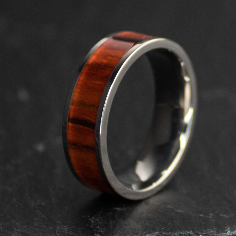 May include: A close-up of a ring with a polished, silver-colored metal band. The ring features a wide, reddish-brown wood inlay. The wood grain is visible, adding texture and warmth to the design. The ring is likely a piece of jewelry.