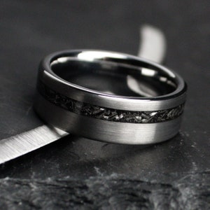 May include: A silver ring with a dark, textured inlay. The ring has a brushed metal finish and a band of dark material. The ring is set against a dark background. The ring's design is modern.