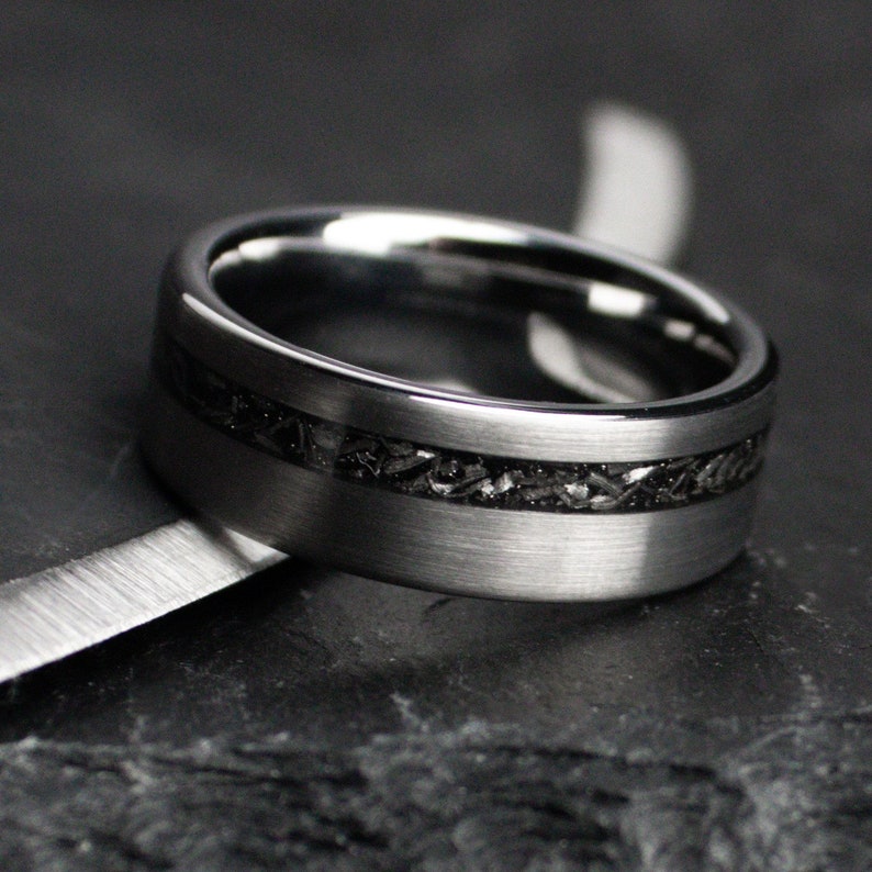 May include: A silver ring with a dark, textured inlay. The ring has a brushed metal finish and a band of dark material. The ring is set against a dark background. The ring's design is modern.