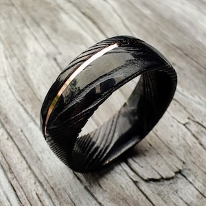 May include: A black ring with a textured pattern and a thin, copper-colored stripe. The ring has a polished, reflective surface. The ring is a piece of jewelry.