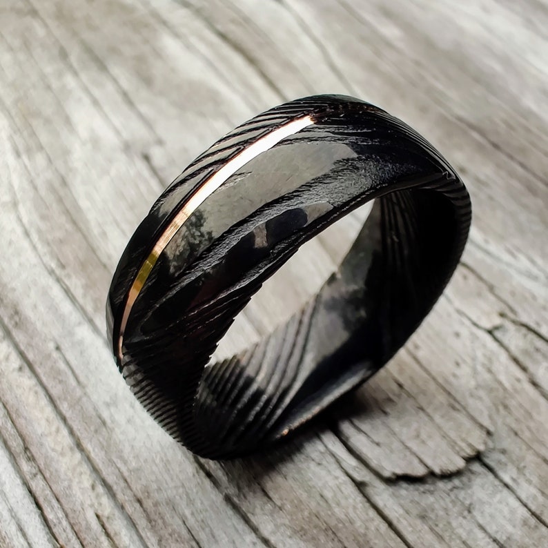 May include: A black ring with a textured pattern and a thin, copper-colored stripe. The ring has a polished, reflective surface. The ring is a piece of jewelry.