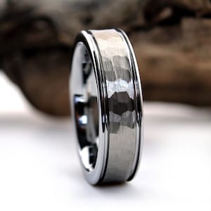 May include: Close-up of a silver ring with a hammered center band. The ring has a polished, shiny finish with a textured, matte center. The ring is a wedding band or fashion accessory.
