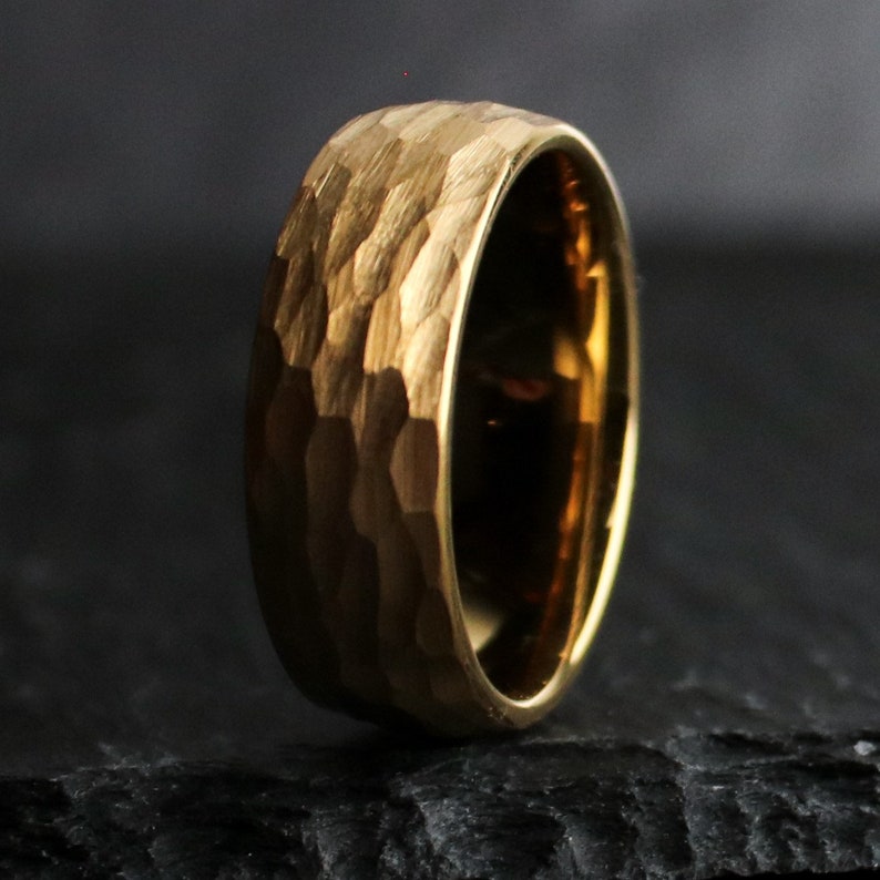 May include: A gold-colored ring with a textured, hammered surface. The ring has a wide band and a polished interior. The ring is displayed against a dark background, highlighting its metallic sheen. The ring's design suggests a handcrafted or artisan style.
